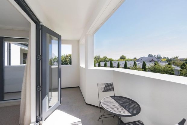 Bright, minimalist covered balcony or terrace with white rendered walls, a dark-framed glazed folding door, smooth grey flooring, and a compact black perforated metal bistro table and chairs. The open parapet frames views of neighbouring homes and clipped greenery beyond.
