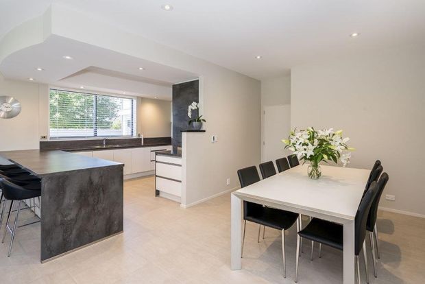 Bright, open-plan contemporary kitchen and dining area with a large dark stone island, white cabinetry, timber-toned flooring, recessed ceiling lighting, and a white dining table surrounded by black chairs. A wide window with blinds brings in natural light, while a dark feature panel and floral arrangement add contrast.