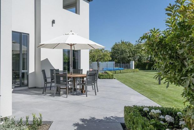 Exterior view of a contemporary white-rendered house opening onto a broad paved terrace. The terrace contains an outdoor dining table and chairs beneath a large white umbrella, with a manicured lawn, clipped hedges, mature planting and a fenced blue swimming pool visible beyond.
