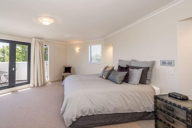 Bright, spacious bedroom with a large upholstered bed, layered neutral and patterned cushions, carpeted flooring, cream walls, and white crown moulding. Full-height glazed doors open to a small balcony, with sheer curtains and additional windows providing abundant natural light. A dark metal-trimmed chest sits beside the bed.