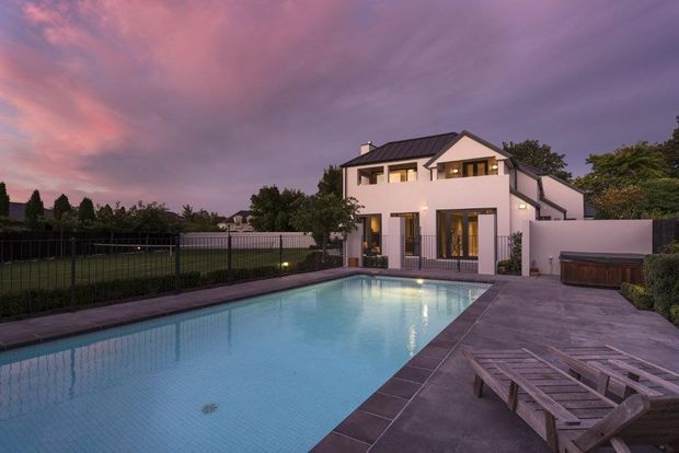 A contemporary two-storey white rendered home viewed from the rear, featuring a large rectangular swimming pool, paved pool terrace, timber sun loungers, dark metal fencing, and landscaped surroundings. The house has a pitched gabled roof with dark roofing, large glazed doors and windows, and warm interior lighting, photographed at dusk beneath a dramatic pink and purple sky.