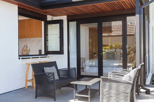 A bright, modern outdoor lounge or patio adjoining a building, furnished with woven black and light-grey armchairs, a small coffee table, and a compact timber console. The space has dark-framed glazed folding or hinged doors, a white-painted brick wall, a timber slatted ceiling, and grey tiled flooring. Reflections reveal an adjacent interior and planted outdoor area.