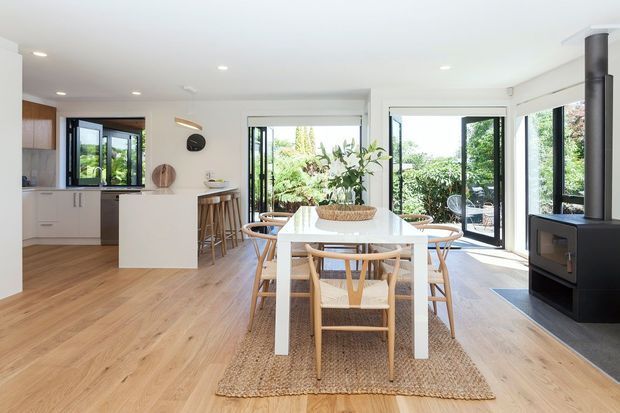 Bright open-plan kitchen and dining area with light timber flooring, white cabinetry and island, a white dining table with timber chairs, and expansive black-framed glazed doors opening to a leafy outdoor space. A modern freestanding black wood-burning stove forms a strong feature at the right.