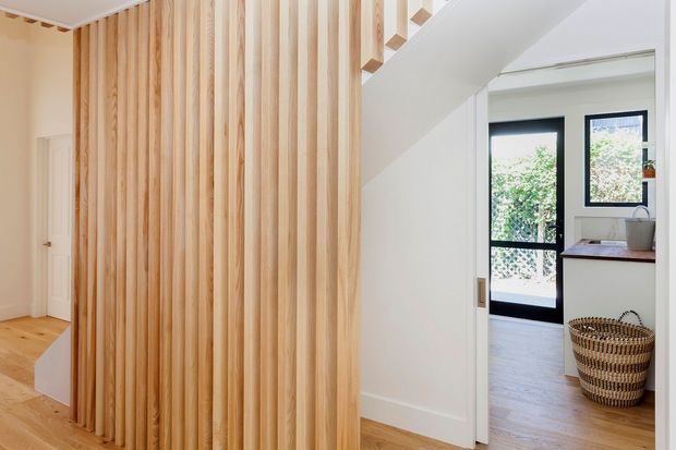 Bright contemporary interior hallway with a dramatic vertical timber-slat feature wall and matching light timber flooring. A white-framed sliding door opens toward a compact kitchen or utility area with dark-framed glazing, timber-toned cabinetry or benchtop, and a woven basket. A white staircase or angled ceiling element adds architectural interest.