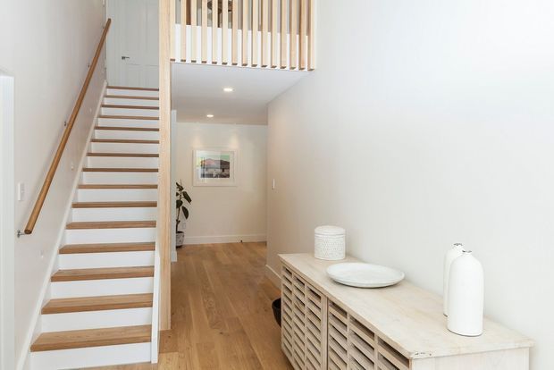 Bright minimalist hallway and stairway interior with white walls, warm timber flooring, an open-riser timber staircase, and a slatted timber balustrade overlooking the entry corridor. A pale timber slatted console with restrained ceramic decor sits along the right wall.