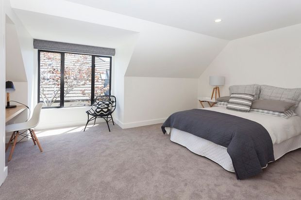 Bright, compact attic-style bedroom with a sloped ceiling, large black-framed window, carpeted floor, and a neatly made bed with grey bedding. A small desk and chair sit beside the window, with a patterned accent chair and bedside lighting adding functional furnishings.