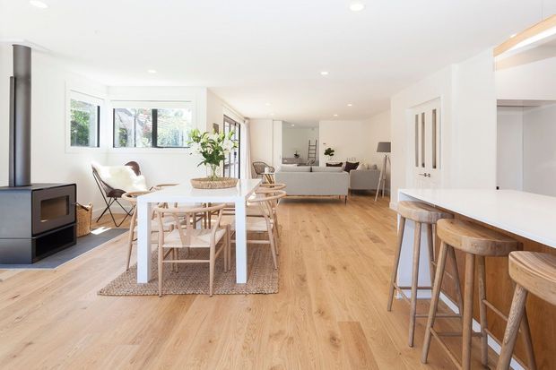 Bright, open-plan contemporary home interior with a combined kitchen, dining and living area. The space features pale timber flooring, white cabinetry and walls, a white kitchen island with timber bar stools, a white dining table with light timber chairs, a black freestanding wood-burning stove, and large windows providing abundant natural light. A lounge area is visible beyond the dining space.