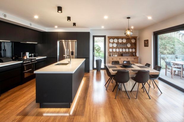 Open-plan kitchen and dining area with a large black island, integrated dark cabinetry, stainless-steel appliances, and a light stone benchtop. Warm timber flooring extends through the room to a timber dining table and built-in plate shelving. Large glazed doors and windows provide abundant natural light, while black track and pendant lighting add contrast.