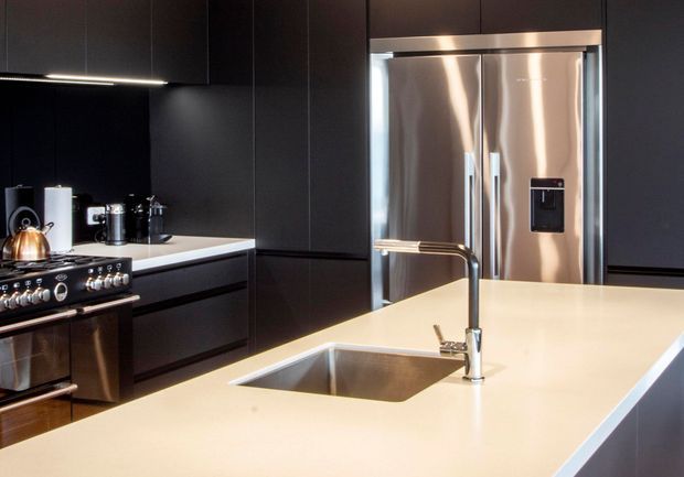 A sleek contemporary kitchen dominated by matte black full-height cabinetry and drawers, with a large pale stone island featuring an integrated stainless-steel sink and streamlined brushed-metal tap. Stainless-steel appliances, including a freestanding range and double-door refrigerator, form the main focal points. Under-cabinet lighting creates a polished, high-contrast look.