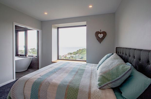 A cozy modern bedroom with a large upholstered button-tufted bed, layered pillows, and a muted striped quilt. A wide picture window provides a coastal ocean view, while an adjacent open doorway reveals a freestanding bathtub and a second window, suggesting a directly connected ensuite bathroom. The room has pale grey walls, recessed ceiling lights, dark carpeting, and warm timber window reveals.