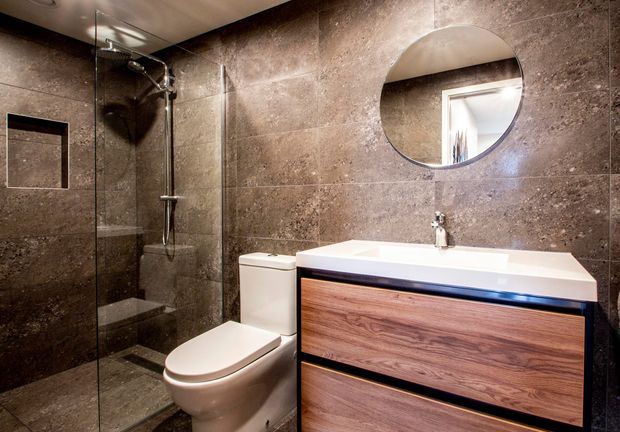 Compact modern bathroom with full-height dark brown stone-look wall and floor tiles, a frameless glass walk-in shower, wall-hung shower fittings, white toilet, round mirror, and a contemporary timber-look floating vanity with white benchtop and integrated basin. The space has a refined, slightly dramatic finish with concealed storage and chrome fixtures.