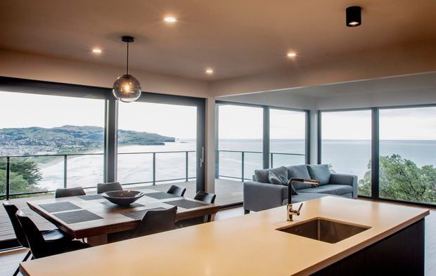 An open-plan contemporary kitchen, dining and living area with expansive corner glazing overlooking a coastal or waterfront landscape. The kitchen features a large pale stone island with integrated sink and dark fixed cabinetry, while the dining area has a substantial timber table and dark upholstered chairs. A grey sofa sits beside the glazing, and slim dark-framed windows and glass balustrades create a bright, elevated indoor-outdoor connection.