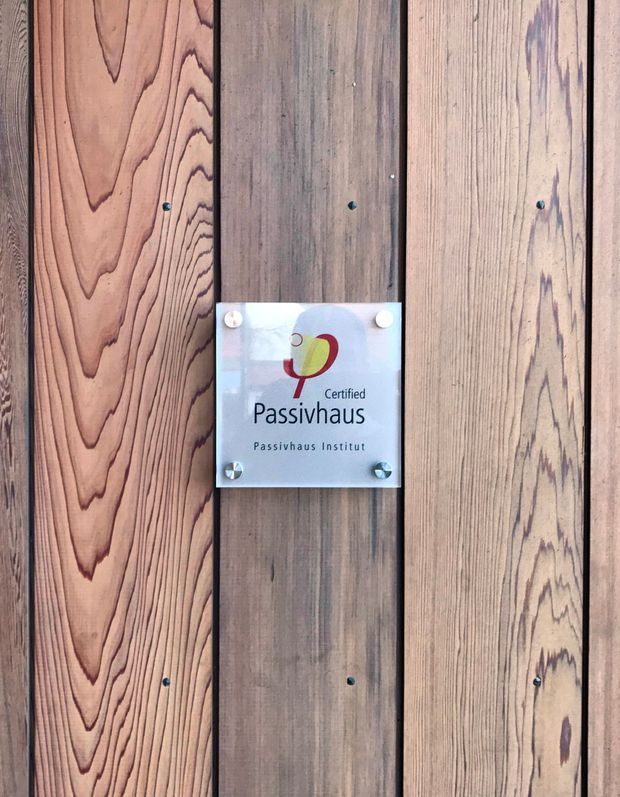 Close-up of a certified Passivhaus plaque mounted on a vertically clad timber wall. The wall consists of contrasting natural timber boards with prominent grain and visible fasteners; the plaque reads “Certified Passivhaus – Passivhaus Institut.”