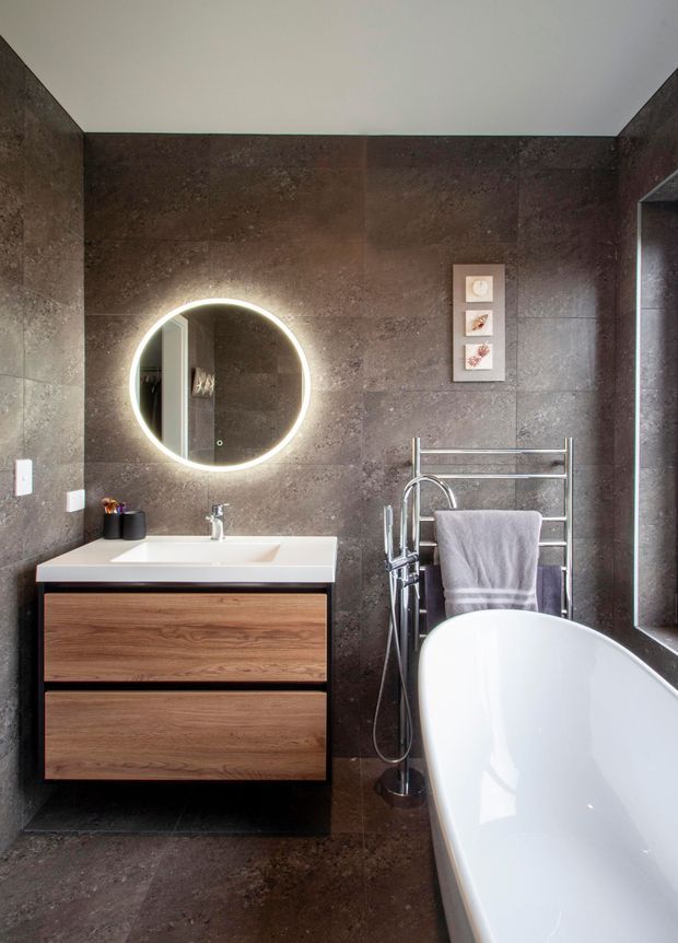A contemporary bathroom with dark brown stone-look wall and floor tiles, a floating timber vanity with a white integrated basin, a backlit round mirror, chrome tapware, heated towel rail, and a freestanding white bathtub. The room has a refined, moody atmosphere with clean modern fixtures.