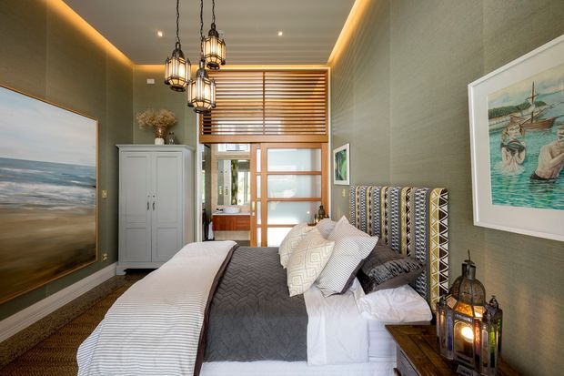 A compact, warmly lit bedroom with a layered bed dressed in white, grey, and patterned textiles. The room features textured green-grey walls, dark timber flooring, a tall upholstered geometric headboard, a white wardrobe, framed artwork, timber Venetian blinds, and a timber-framed glazed doorway. Multiple hanging lantern-style pendant lights and concealed ceiling lighting create a refined, cozy atmosphere.