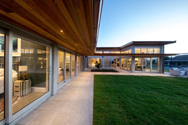 Contemporary single-storey courtyard residence arranged around a manicured central lawn. The image looks along a covered outdoor walkway with extensive floor-to-ceiling glazing and a warm timber soffit, toward a low flat-roofed wing with timber and glazed detailing. A paved terrace and outdoor seating area are visible to the right, with the interior spaces seen through the windows.