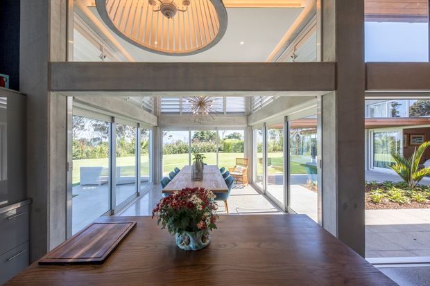 A bright, high-ceilinged dining and living space viewed across a large timber table. Full-height glazing opens the room to a landscaped garden and poolside terrace. A dramatic oval ceiling feature with timber slats and dark trim is visible above, along with exposed-looking structural framing and clerestory-style glazing. The interior combines warm timber, pale walls, expansive glass, and contemporary furnishings.