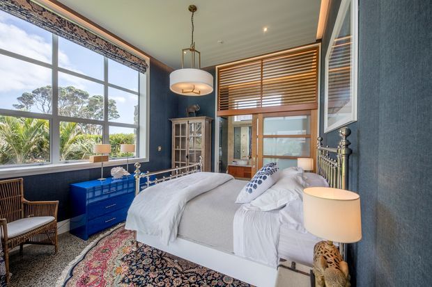 A richly styled bedroom with a white upholstered bed, layered white bedding, dark navy-blue textured walls, and large multi-pane windows. The room includes a vivid blue chest of drawers, a glass-front cabinet, timber-framed glazed doors with horizontal timber blinds, ornate metal bed detailing, patterned rug, wicker seating, and pendant and bedside lighting. The overall look is traditional with eclectic, refined furnishings and strong colour contrast.