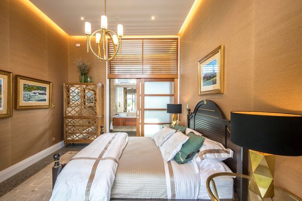Warmly styled bedroom with a large upholstered bed, layered white bedding, dark curved headboard, bedside lamps, framed artwork, and a decorative wood cabinet. The room features textured brown walls, recessed ceiling lighting with concealed warm LED strips, timber-look flooring, and a rear window with horizontal timber blinds and a frosted-glass door.