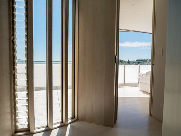 A bright, minimalist interior hallway or entry area with tall vertical timber-framed glazing and a louvred window on the left. A partially open timber door leads toward a bedroom or living space with a bed or upholstered seating visible. Beyond the glazing is a sunlit terrace or balcony overlooking blue water and a distant shoreline. The interior uses pale timber wall panels, light flooring, and clean contemporary detailing.