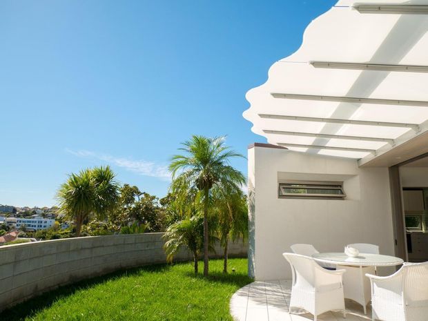 Bright exterior patio or terrace beside a white modern building, featuring a scalloped translucent roof canopy, white outdoor dining furniture, tiled paving, a rendered wall, and a landscaped lawn with palm trees. A suburban neighbourhood is visible beyond the low masonry boundary wall.