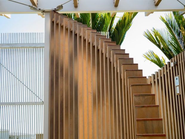 Close-up architectural view of an outdoor timber staircase and vertical timber screening beneath a light-colored pergola or canopy. The stair stringer and repeated vertical battens create a strong geometric rhythm, with palm foliage visible beyond and a bright open-air setting.