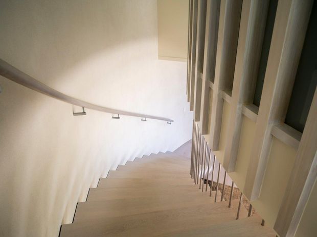 A contemporary stairway viewed from above, with pale timber treads descending alongside a smooth light wall. A slim metal handrail runs along the wall, while a vertical balustrade and framed openings create a strong geometric rhythm on the right. The palette is light and neutral with soft natural light.