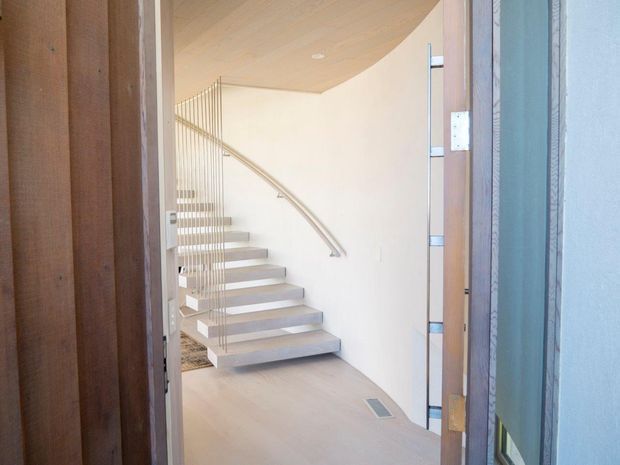 View through an open doorway into a bright, minimalist stairway. The space features a gently curving staircase with pale treads, slender vertical metal balusters, a smooth curved handrail, light timber flooring and ceiling, and white sculpted walls.