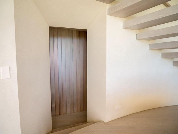 Minimal interior passage or stair landing with warm white plaster walls, pale timber flooring, and a dark vertical timber door set into a recessed opening. Exposed light timber ceiling beams project horizontally across the upper right, creating a strong linear architectural feature. The space is sparsely furnished and softly lit.