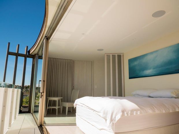Bright contemporary bedroom with a low-profile bed, white bedding, pale carpet, built-in wardrobe joinery, and a small seating area. Floor-to-ceiling curved glazing opens onto a narrow balcony or terrace, bringing in abundant natural light. The room has a clean white palette with a large blue artwork and slim dark exterior framing.