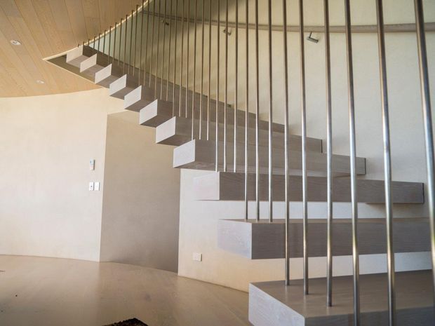 Minimalist contemporary interior featuring a sculptural floating staircase with pale solid treads, dark horizontal riser or stair-edge detailing, and a slender vertical metal balustrade. The staircase rises along a light plaster wall beneath a warm timber-lined ceiling, with a pale smooth floor and restrained architectural detailing.