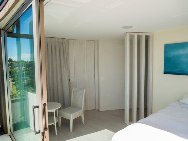 Bright contemporary bedroom with a white bed, pale neutral walls, light flooring, and a compact sitting area by full-height glazed doors. A partially screened wardrobe or room divider is visible, with a blue artwork panel on the wall and an exterior landscape seen through the glazing.