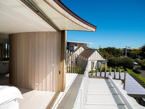 Bright architectural view from an upper-level outdoor terrace or balcony, looking along a contemporary home with pale vertical timber wall cladding, white decking or paving, a curved projecting roof edge, and white balustrades. A bedroom is partially visible at left, while neighbouring pitched-roof homes and suburban streets appear beyond the terrace.