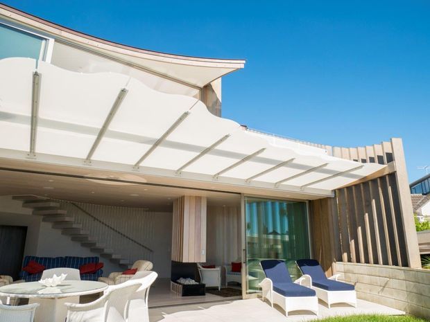 Modern multi-level residential exterior with a covered outdoor entertaining area and patio. The scene features a translucent white tensile-style canopy, outdoor dining furniture, lounge chairs, a glazed sliding door, vertical timber-look screening, a sculptural open stair, and a bright coastal atmosphere.