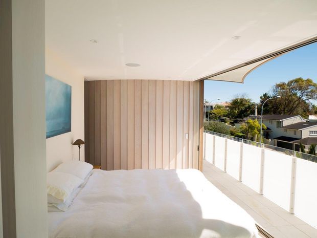 Bright contemporary bedroom opening onto a spacious balcony or terrace. The room features a white bed, pale timber vertical wall panelling, recessed ceiling lighting, and large open glazing with a frameless glass balustrade overlooking a suburban setting.