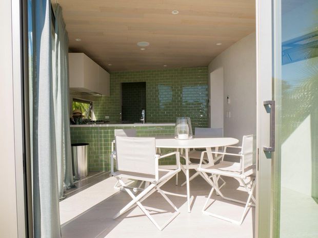 Bright compact open-plan kitchen and dining area viewed through open glazed doors. The space features a green glazed-tile kitchen backsplash and island front, white built-in cabinetry, a warm timber-lined ceiling, pale flooring, and a white outdoor-style dining table with folding chairs. Strong daylight and garden reflections create an airy indoor-outdoor feel.