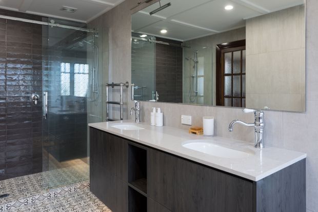 Contemporary bathroom featuring a spacious double vanity with a white stone benchtop, dark timber-look cabinetry, large wall mirror, chrome fixtures, a glass-enclosed shower, and patterned encaustic-style floor tiles. Neutral grey and beige wall finishes create a refined, practical interior.
