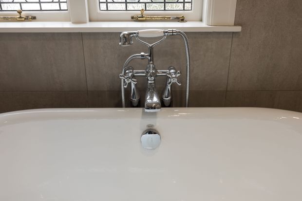 Close-up view of a freestanding white bathtub with polished chrome traditional-style taps, handheld shower, and exposed plumbing fixtures. The bath is set against large-format taupe-grey wall tiles beneath leaded or textured glass windows with white-painted frames.