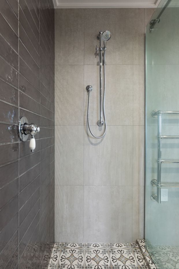 A compact shower area in a bathroom, featuring large light beige wall tiles, dark glossy rectangular tiles on the side wall, a handheld chrome shower fixture, patterned decorative floor tiles, and a clear glass shower screen. A chrome heated towel rail is visible behind the glass.