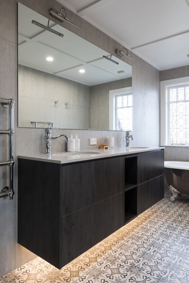 A refined bathroom with a large wall-to-wall mirror, double integrated vanity basins, dark timber-look cabinetry, chrome wall-mounted taps, and a pale stone or solid-surface benchtop. Decorative patterned floor tiles add visual interest, while a freestanding bath and ornate leaded-style window are visible on the right. The room combines contemporary joinery with traditional architectural detailing.