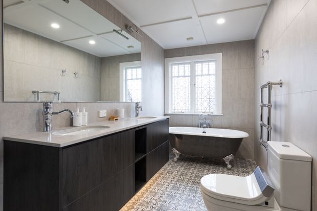 A refined traditional bathroom with a freestanding clawfoot bathtub beneath a leaded-glass window, a wall-mounted heated towel rail, toilet, and a long dark timber vanity with integrated white basins. Large pale wall tiles, a mirrored vanity wall, patterned encaustic-style floor tiles, and chrome fixtures create a bright, elegant scheme.