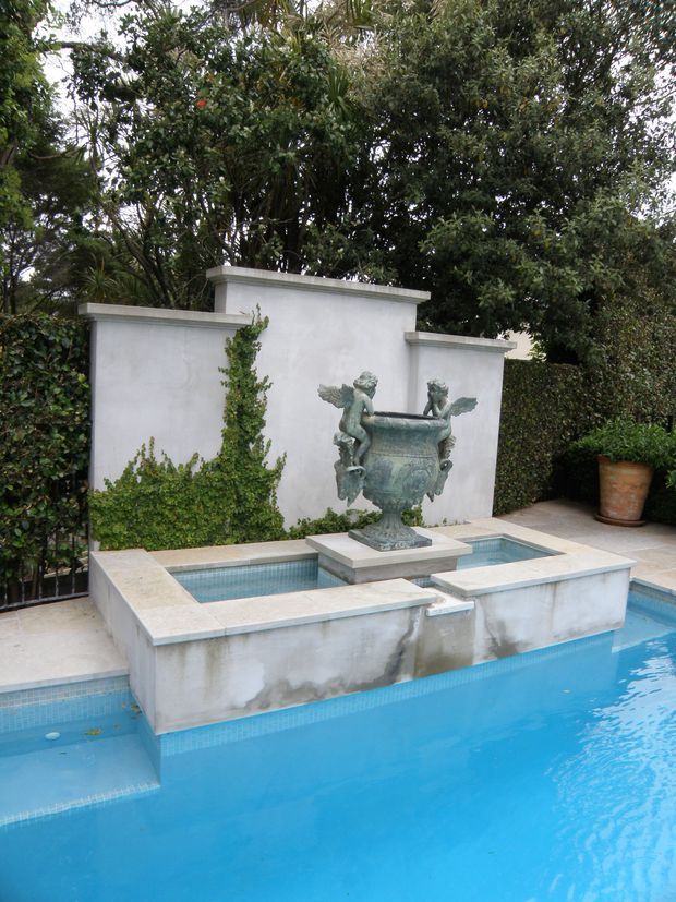 A formal landscaped courtyard pool with a raised rectangular fountain basin and central ornate urn sculpture featuring winged figures. The fountain is set against a pale rendered garden wall with clipped hedging, dense mature planting, terracotta planter pots, and pale stone coping surrounding bright blue tiled water. The composition has a classical, refined garden character.