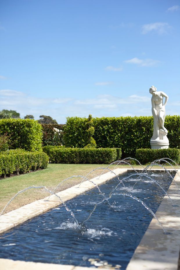 A formal landscaped garden featuring a long rectangular reflecting pool with arcing water jets, pale stone edging, neatly clipped box hedges, and a classical white female statue. The scene is brightly lit under a clear blue sky.