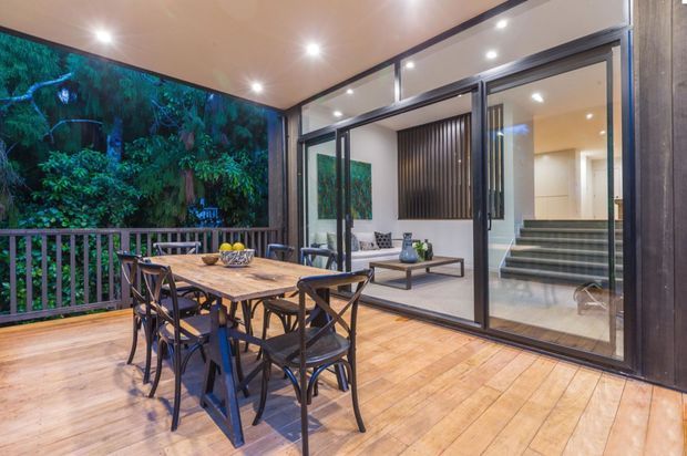 Covered timber deck dining area opening into a contemporary living space through large black-framed sliding glass doors. The deck includes a timber dining table with black metal chairs, while the interior shows a sofa, coffee table, stairs, and vertical feature screening. Dense greenery is visible beyond the deck railing.
