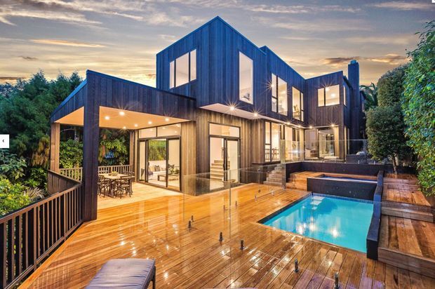 Contemporary multi-level residence clad in dark vertical timber, with extensive floor-to-ceiling glazing opening onto a large timber deck. The deck includes a bright swimming pool, integrated spa-like seating, outdoor dining, lush planting, and warm recessed lighting, photographed at dusk after rain.
