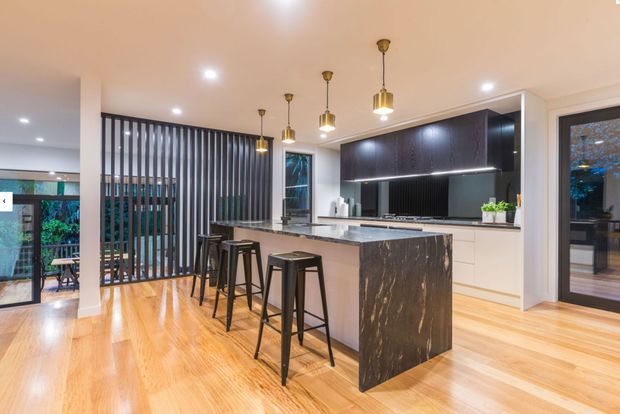 Contemporary open-plan kitchen with a large stone island and waterfall edge, four black stools, dark timber cabinetry, white lower cabinets, pendant lighting, warm timber flooring, and a vertical slatted room divider. Glazing and a glazed door provide views to greenery outside.