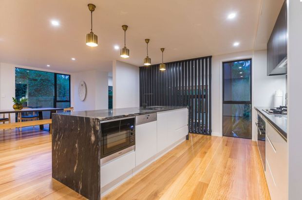 Contemporary open-plan kitchen with a large dark stone island, white flat-panel cabinetry, integrated oven, and timber flooring. Pendant lights hang above the island, while a black vertical slatted screen separates the kitchen from the dining area. Full-height glazing and a glazed external door bring in natural light and views of greenery.