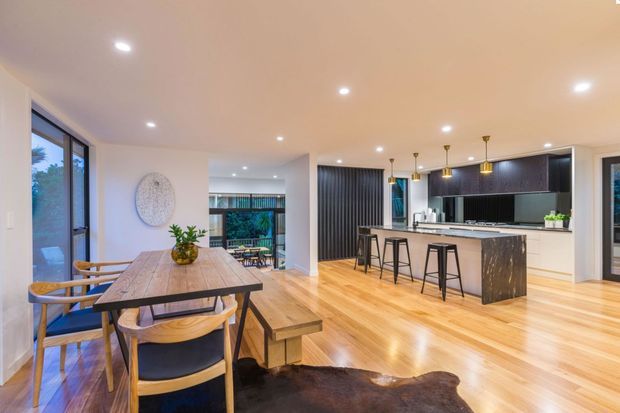 Open-plan contemporary kitchen and dining area with warm timber flooring, a large marble-look island with black bar stools, dark linear cabinetry, pendant lighting, and a timber dining table beside floor-to-ceiling glazing. A living or outdoor area is visible beyond the central opening.