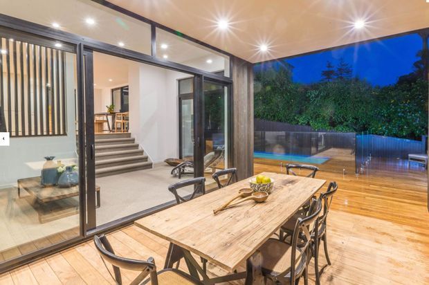 A covered outdoor dining area with a large rustic timber dining table and metal-framed chairs, opening onto a timber deck and landscaped backyard with a pool. Extensive black-framed glass sliding doors connect the space to a contemporary interior, while recessed ceiling lights create a warm evening atmosphere.