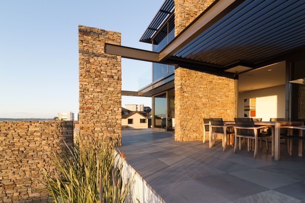 Exterior view of a contemporary multi-level residence with broad stone-clad walls, dark horizontal roof and pergola structures, extensive glazing, and a covered outdoor dining terrace. The terrace overlooks a low stone boundary wall and distant homes, with architectural planting in the foreground.